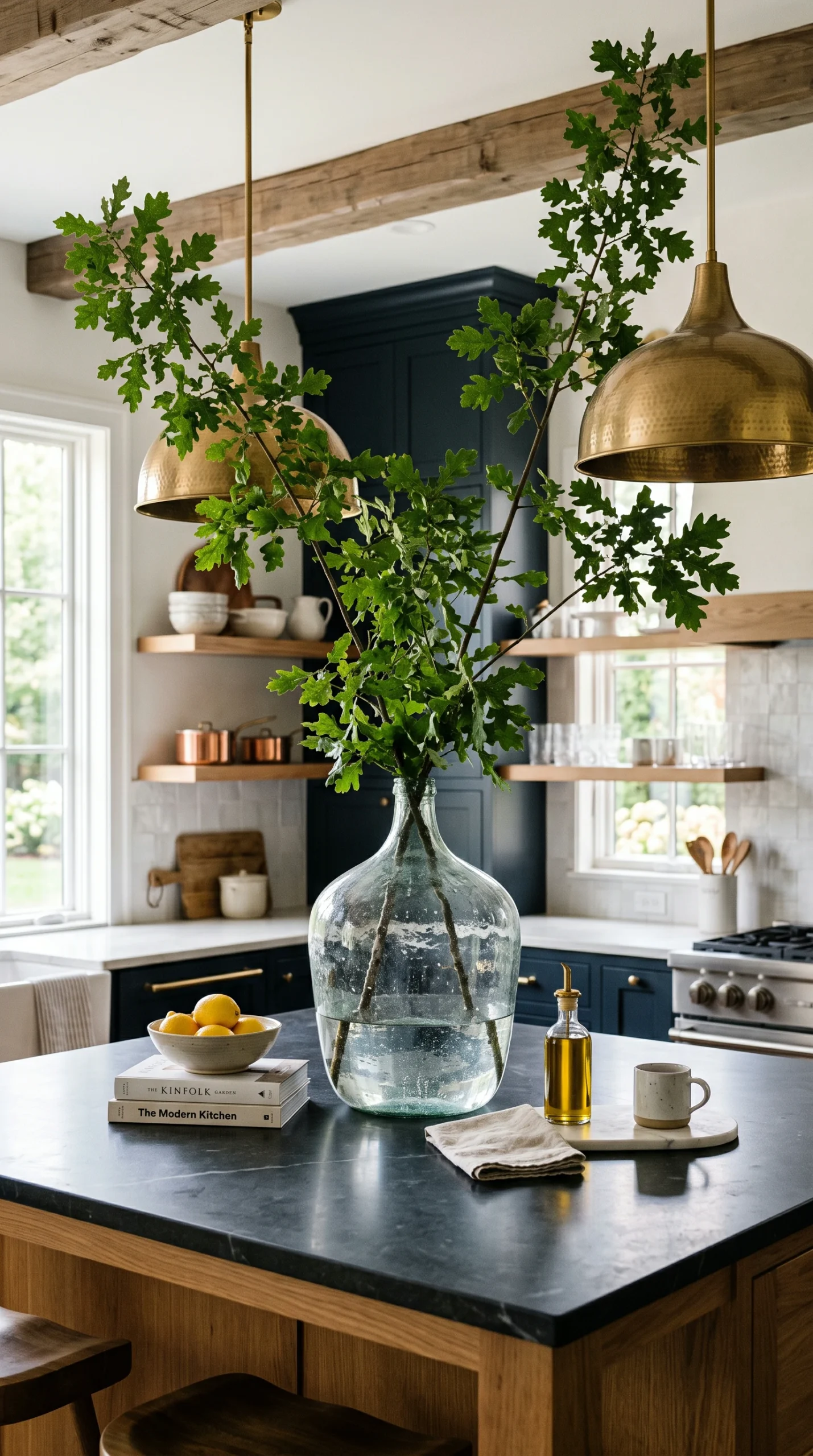 a massive antique glass demijohn vase holding three sprawling four-foot-tall leafy oak branches reaching up toward pendant lighting on a kitchen island.